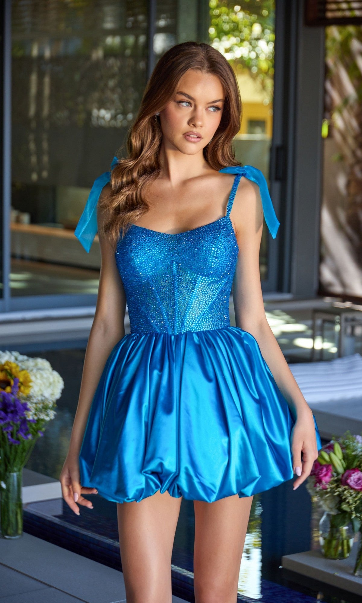 A woman stands by an indoor pool with flowers in the background, wearing the Ava Presley 42386 short homecoming dress featuring a sparkly blue beaded corset bodice and a shiny, voluminous bubble skirt.